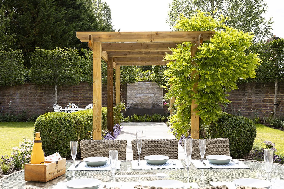 Summer garden ideas, an elegant outdoor dining table set with champagne glasses and plates under a wooden pergola in a landscaped garden.