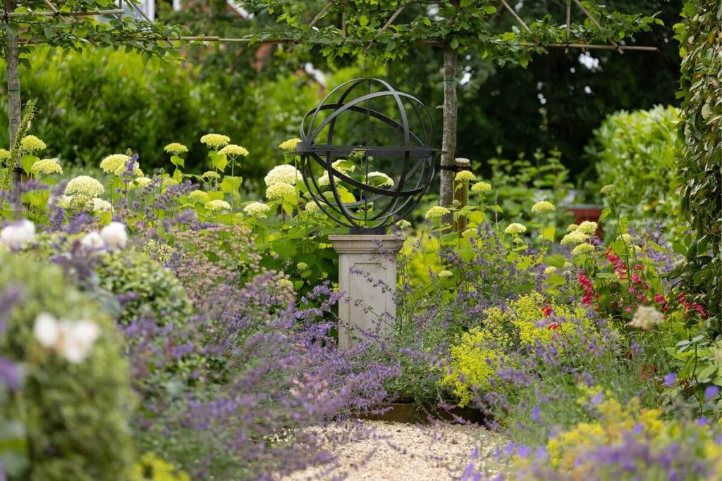 A steel globe on a stone pedestal surrounded by nepeta and salvia flowers in garden beds.