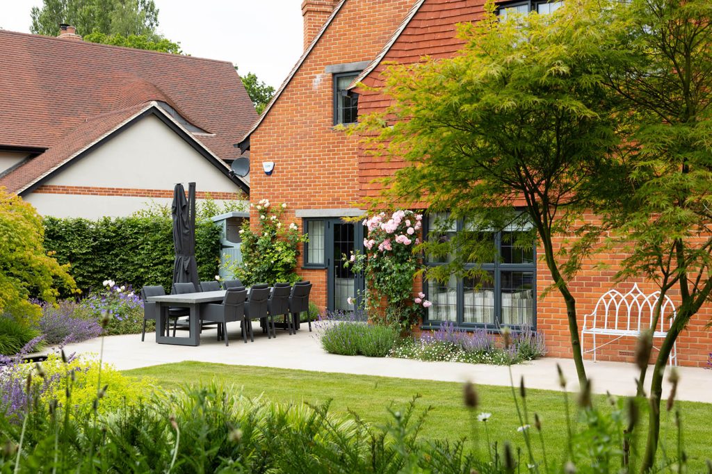 An outdoor patio area features a large rectangular table with grey cushioned chairs. A modern red brick house is in the background with climbing pink flowers. The foreground is filled with lush green plants and a white wrought iron bench is to the right.